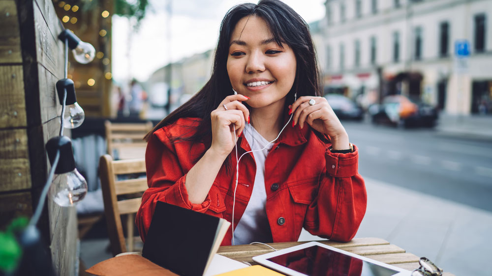 Žena sediaca pri stolíku na ulici s knihami, okuliarmi a slúchadlami pripojenými k tabletu_03