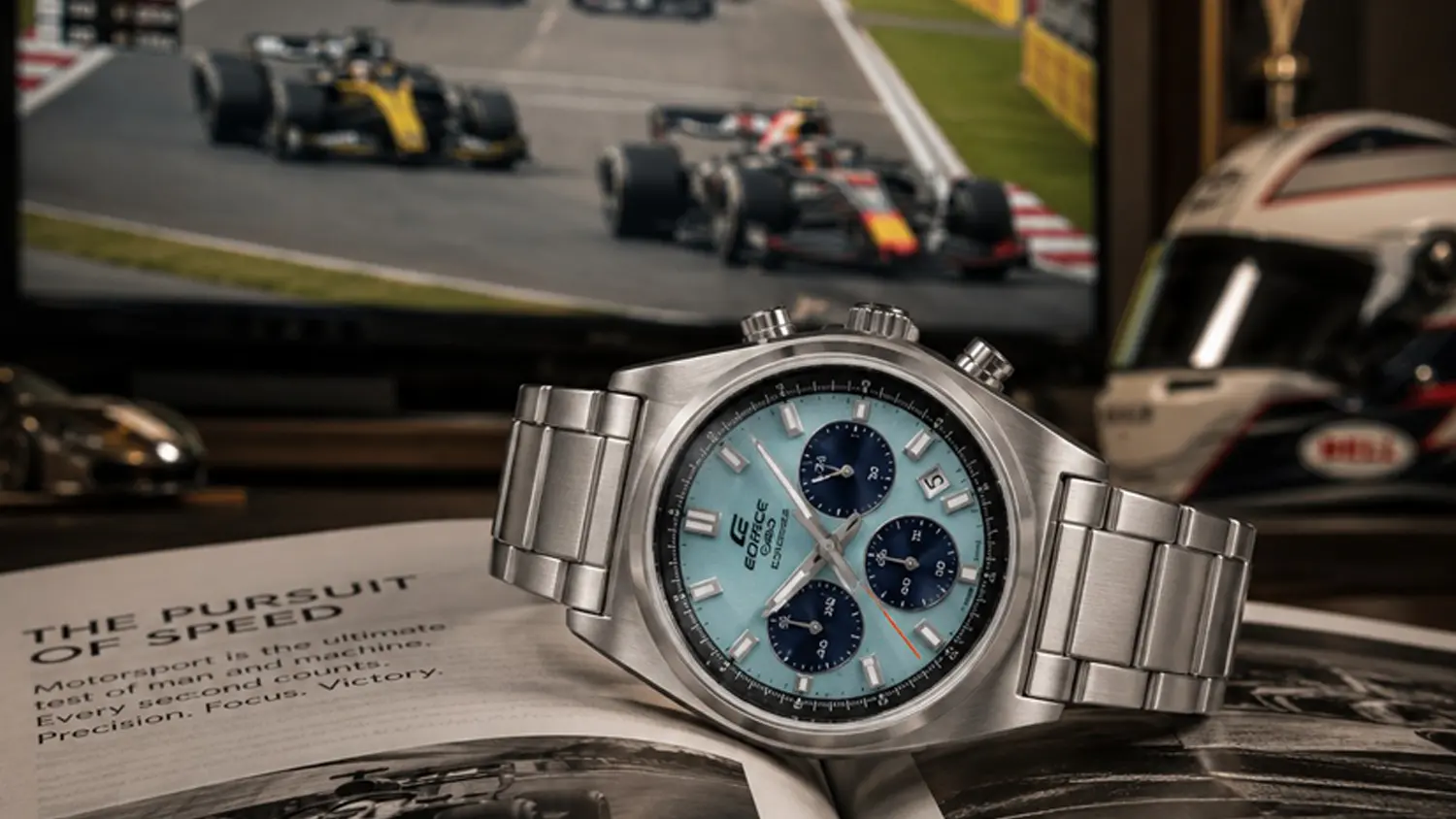 Náramkové hodinky Casio EFB-730D-2BVUEF položené na knize se závody v TV v pozadí