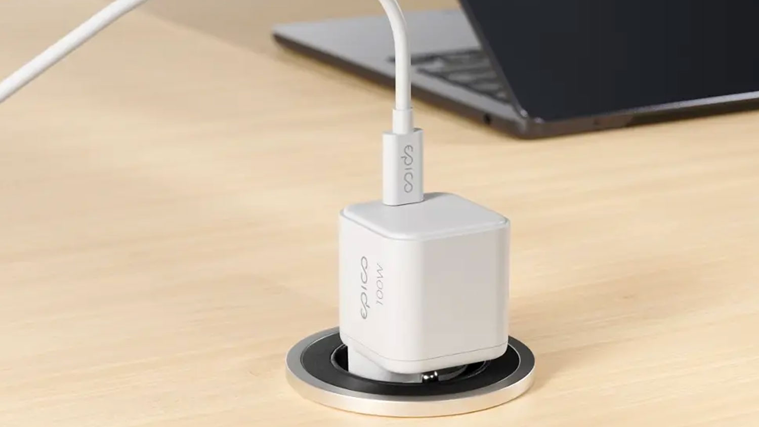 Bílý nabíjecí adaptér Epico 100 W zapojený do stolní zásuvky s připojeným kabelem USB-C a notebookem v pozadí.
