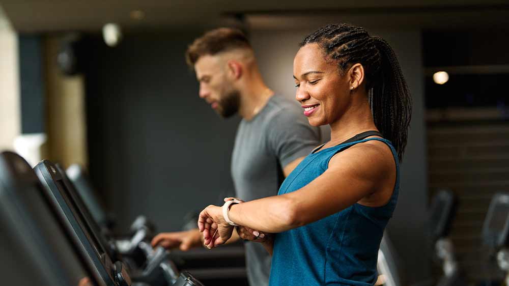 Žena na běžeckém pásu ve fitku kontrolující údaje na chytrých hodinkách na zápěstí a vedle ní muž na běžícím pásu_03