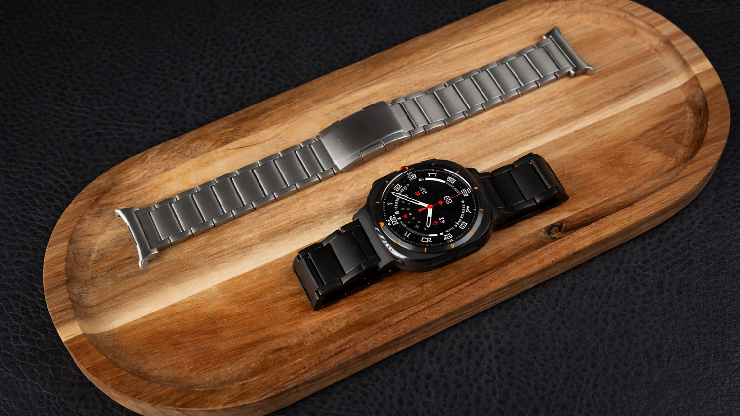 Titanov&yacute; řem&iacute;nek na dřevěn&eacute;m prk&eacute;nku a vedle čern&eacute; hodinky Samsung Galaxy Watch Ultra