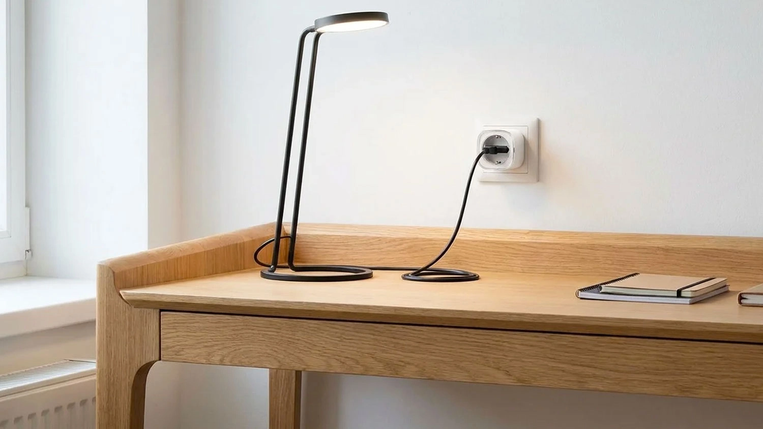 Smart zásuvka EZVIZ T31-16E zapojená ve zdi nad dřevěným stolem napájí rozsvícenou černou stolní lampu.