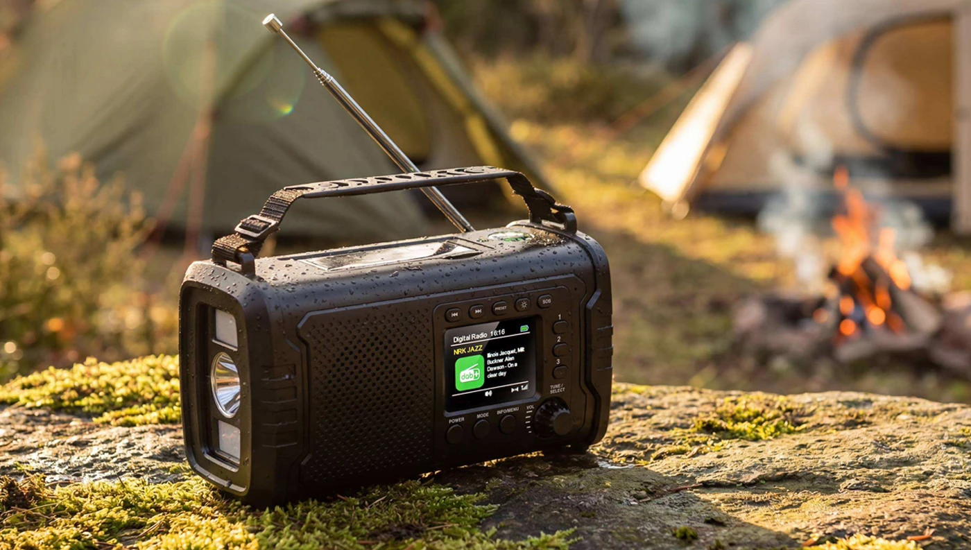 Outdoorové rádio SENCOR SRD 1090 s anténou na skale, v pozadí stany a táborák