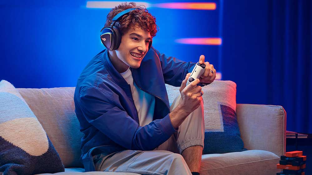 Mladý muž hraje hru na herní konzoli, v ruce má ovladač a má nasazený herní headset ROG Delta II_03