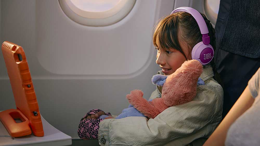 Mladá dívka sedící v letadle a sledující tablet s nasazenými dětskými sluchátky JBL JR320BT_04