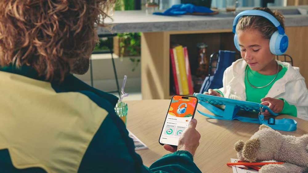Dívka má nasazená dětská bezdrátová sluchátka JBL Junior JR470NC v modré variantě, zatímco rodič provádí nastavení v aplikaci v telefonu_02