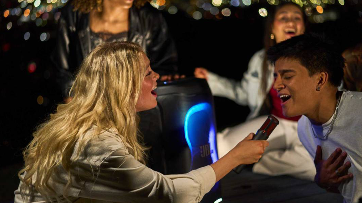 Lidé zpívají do mikrofonu před reproduktorem JBL PartyBox