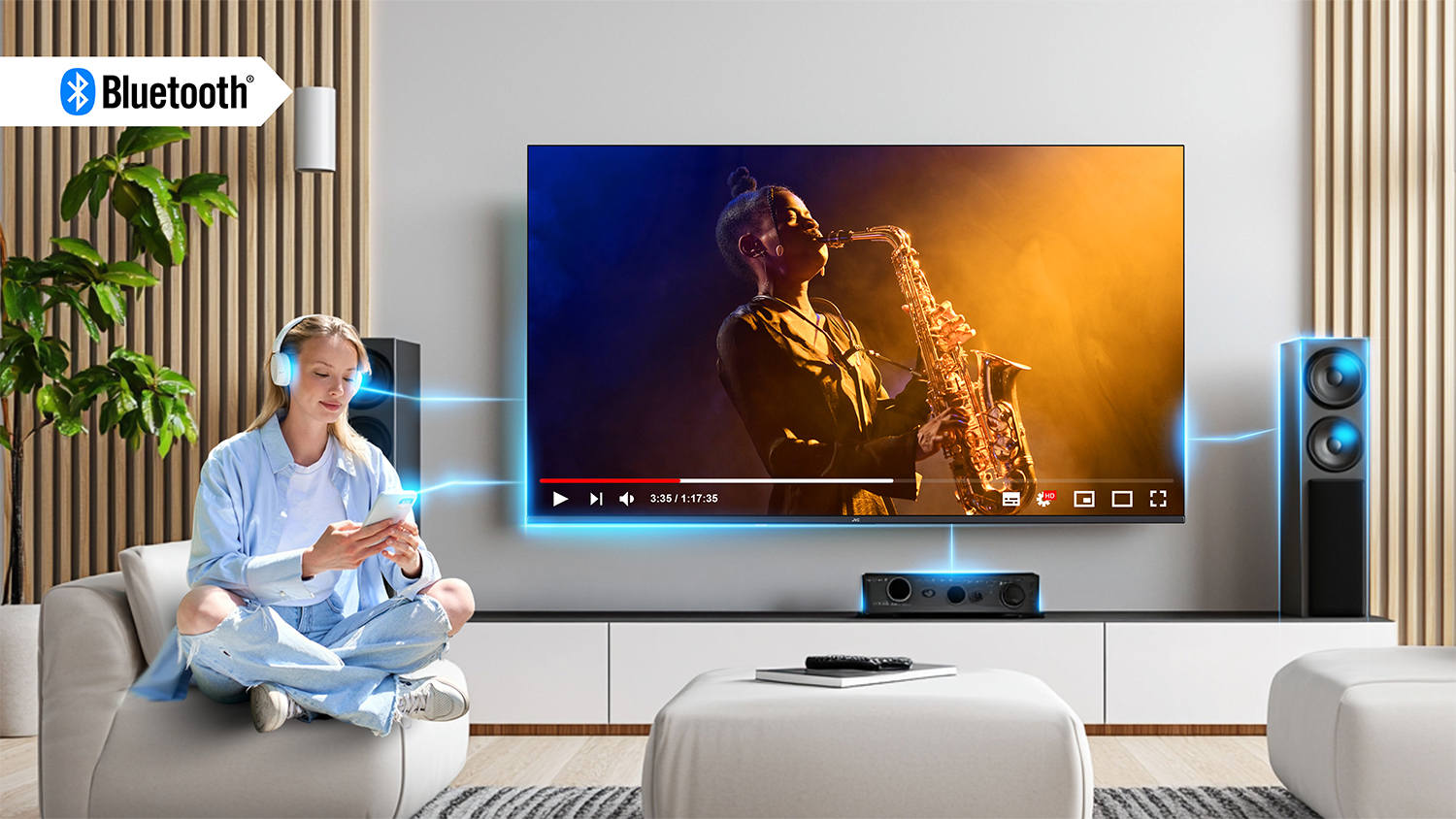 Žena sed&iacute;c&iacute; v ob&yacute;v&aacute;ku poslouch&aacute; televizor JVC se sluch&aacute;tky na hlavě propojena pomoc&iacute; Bluetooth
