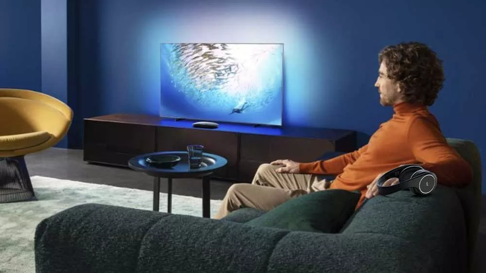 Muž sleduje televizi a m&aacute; vedle sebe připraven&aacute; bezdr&aacute;tov&aacute; sluch&aacute;tka Meliconi HP Digital Smart, zat&iacute;mco z&aacute;kladna vys&iacute;lače je um&iacute;stěna před televiz&iacute;_03
