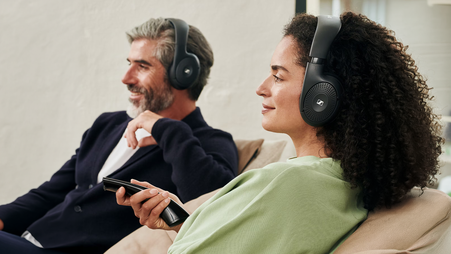 Žena s ovl&aacute;dačom a muž na pohovke so sl&uacute;chadlami Sennheiser RS 120-W