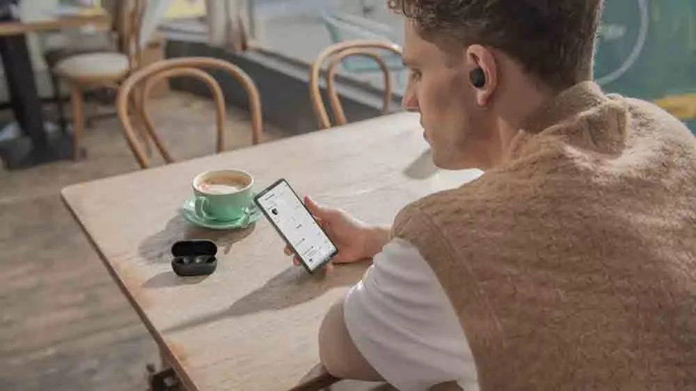 Muž sed&iacute;c&iacute; u stolu drž&iacute; v ruce telefon a v u&scaron;&iacute;ch m&aacute; bezdr&aacute;tov&aacute; sluch&aacute;tka Sony WF-C710N Black_03