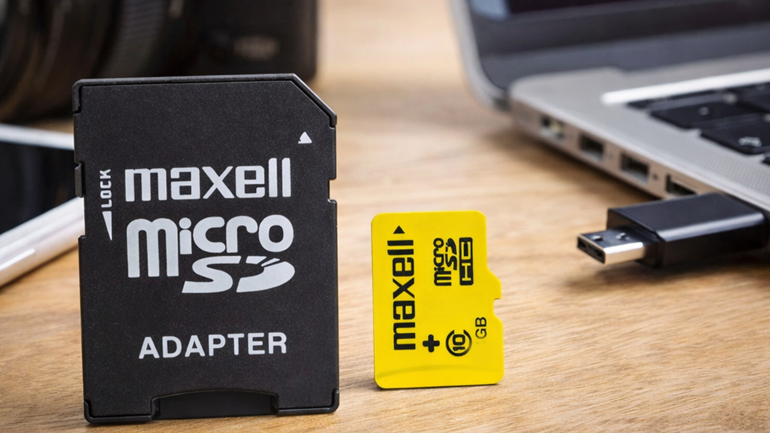 Paměťov&aacute; karta Maxell MicroSDHC 16GB CL10 + Adapt&eacute;r yellow na stole vedle notebooku a fotoapar&aacute;tu