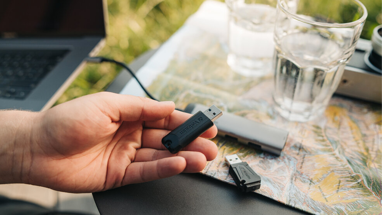 Ruka drž&iacute;c&iacute; USB flash Verbatim, v pozad&iacute; poč&iacute;tač, fotoapar&aacute;t a skleničky_02