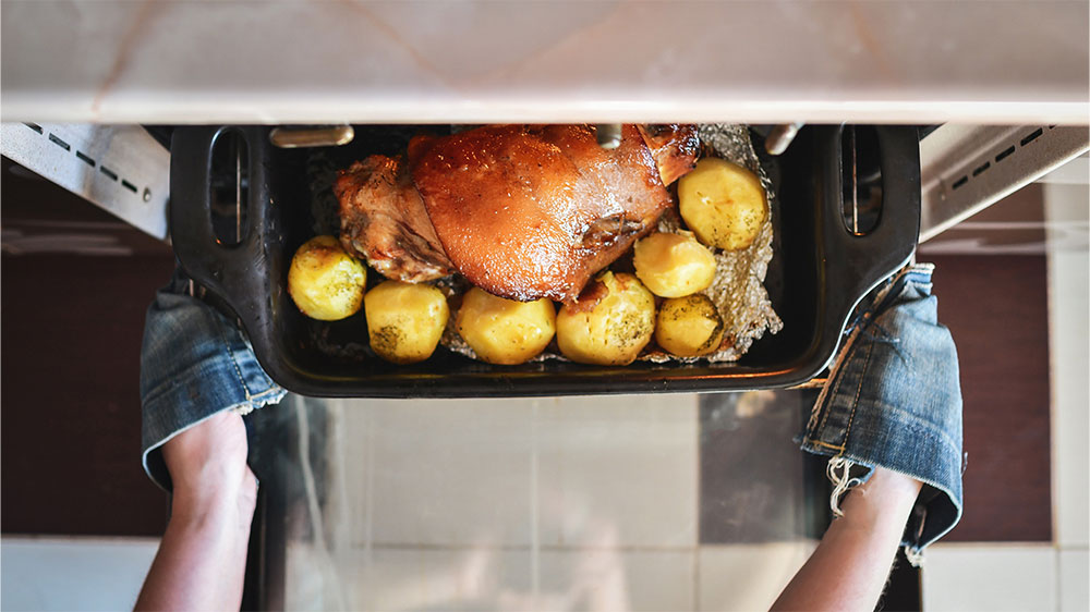 Ruce v chňapkách vyndávají z trouby volně stojícího sporáku Amica SS 19 EAW pekáč s dokonale upečeným vepřovým kolenem s bramborami_04