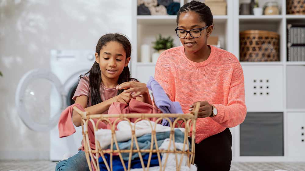 Matka s dcerou klečící na zemi a skládající prádlo z ratanového košíku, za nimi otevřená pračka a policový regál s košíky_04