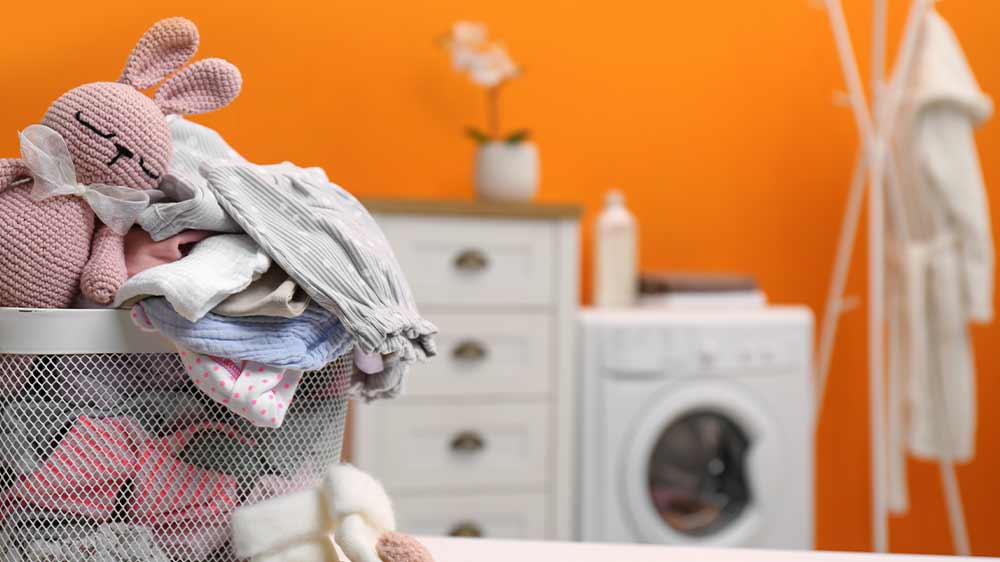 Kovový košík plný dětského oblečení s háčkovaným zajíčkem v levém rohu a v pozadí bílá šuplíková komoda s pračkou a věšákem na oblečení_06