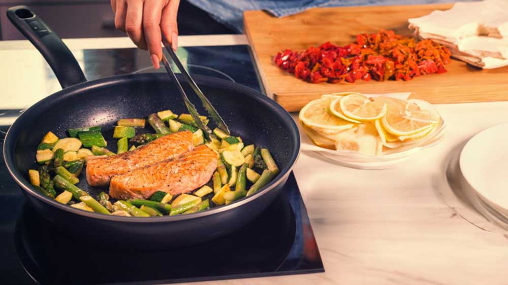 Žena smaží rybu a zeleninu v pánvi na sklokeramické varné desce_03