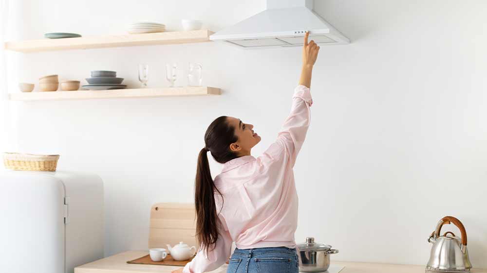 Žena ovládající komínovou digestoř v bílé barvě zavěšenou na zdi nad sporákem v kuchyni_04