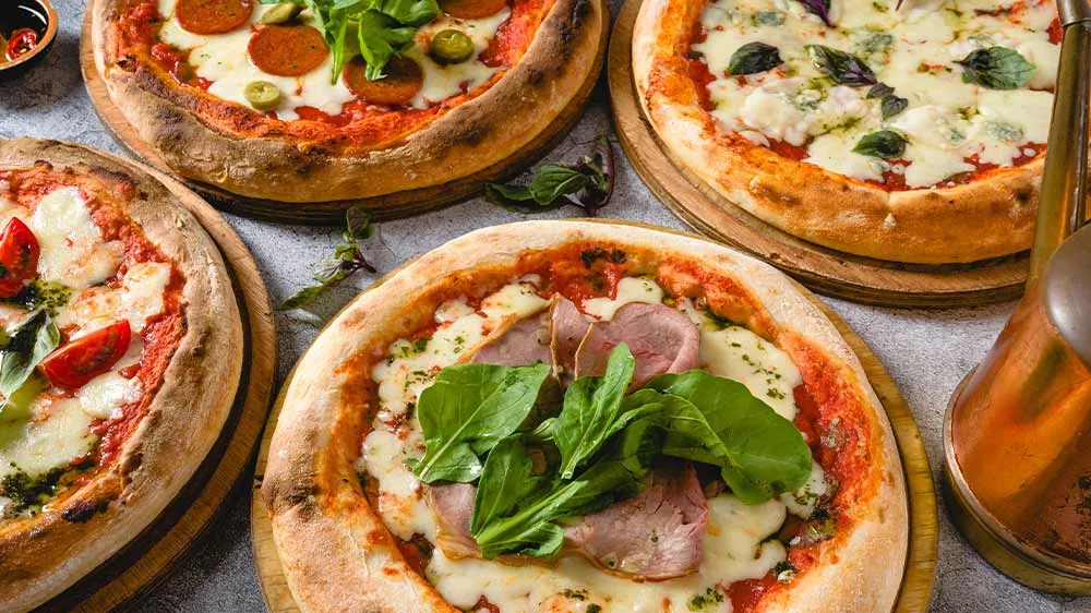 Čtyři druhy pizzy na dřevěných prkénkách, ozdobené saláty, bylinkami a rajčaty s konvičkou na olej na pravé straně_06