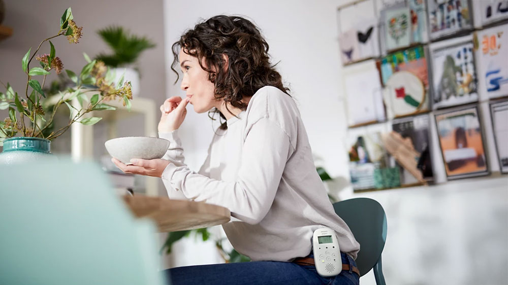 Žena sedící u stolu s miskou jídla a za páskem u kalhot zavěšená audio dětská chůvička Philips Avent Advanced SCD 715/52_05