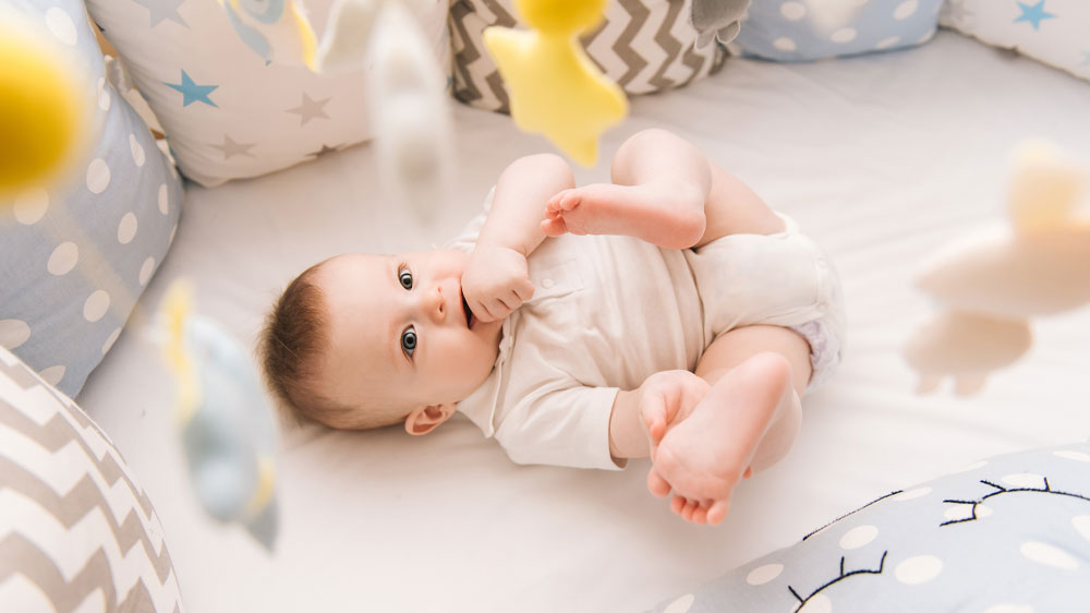 Batole v postýlce s polštářkovými mantinely kolem sebe hrající si s nohama a rukama_05