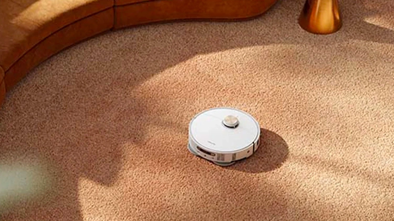Biely robotický vysávač Dreame pri upratovaní béžového koberca vedľa hnedej pohovky.