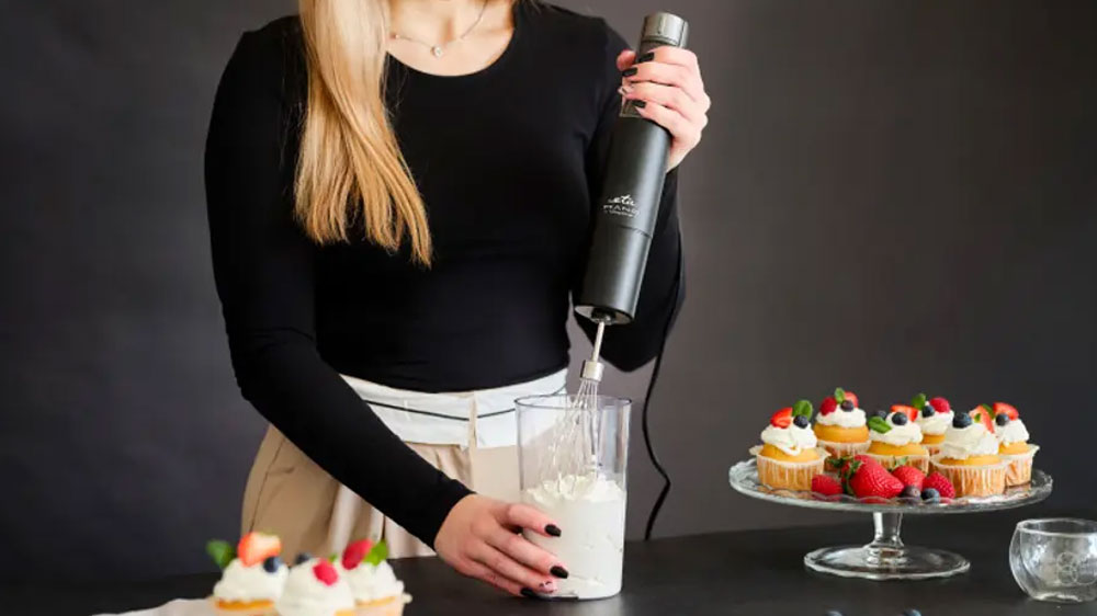 Žena šlehající šlehačku v nádobě tyčovým mixérem ETA Mano 3219 90010 na pracovní desce mezi podnosy s mufiny_02
