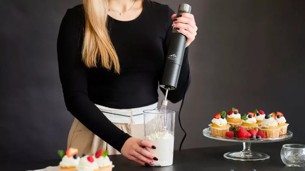 Žena šľahajúcu šľahačku v nádobe tyčovým mixérom ETA Mano 3219 90010 na pracovnej doske medzi podnosy s mufinmi _02