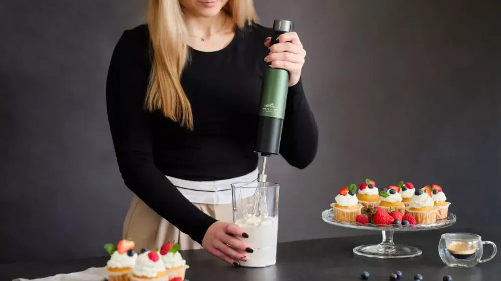 Žena šľahajúcu šľahačku v nádobe tyčovým mixérom ETA Mano 3219 90030 na pracovnej doske medzi podnosy s mufinmi _02