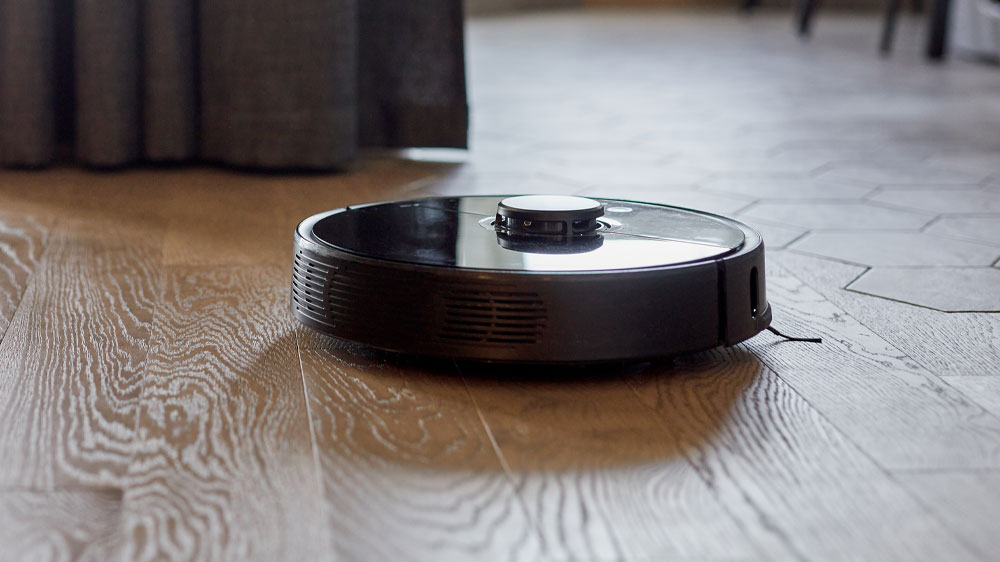 Robotický vysavač čistící plovoucí podlahu a v levé části bosé nohy opřené o zem_02