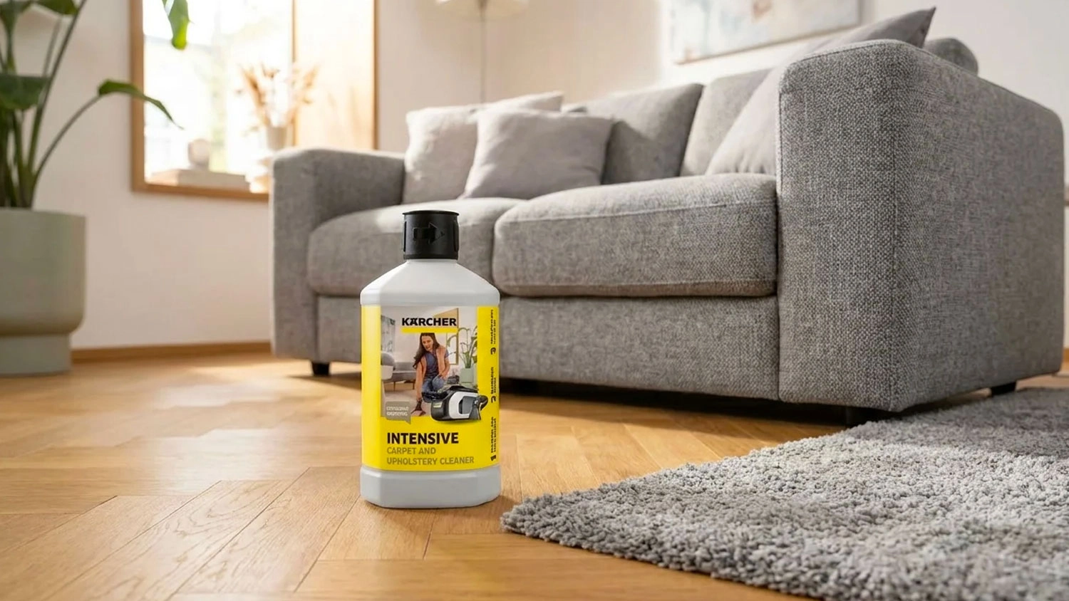 Láhev čističe Kärcher Intensive Carpet & Upholstery Cleaner stojí na dřevěné podlaze vedle koberce a šedé pohovky.