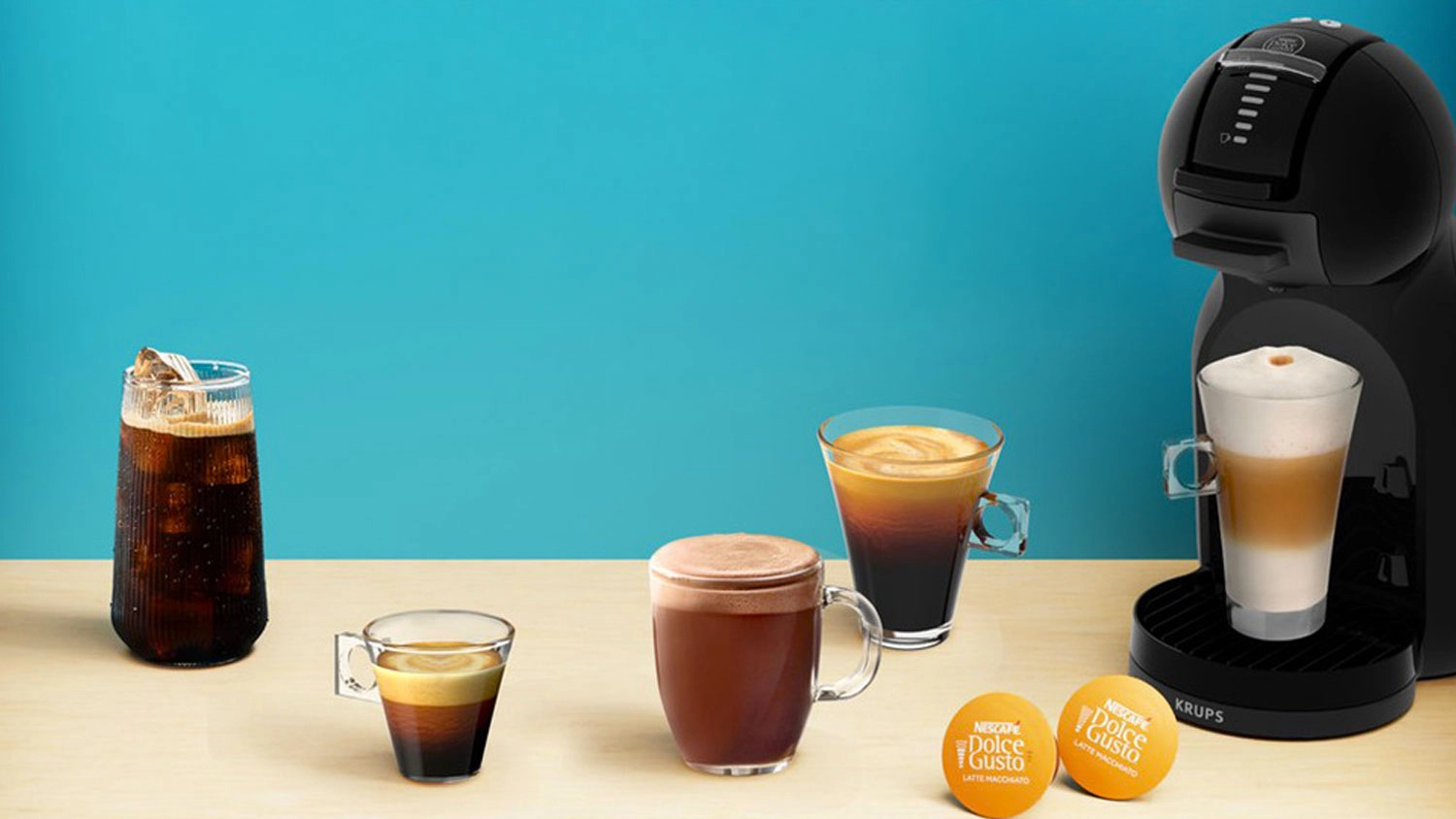 Kávovar na kapsuly Krups Mini Me 2 NESCAFÉ Dolce Gusto vedľa pohára espressa, ľadovej kávy a ďalších šálok kávy