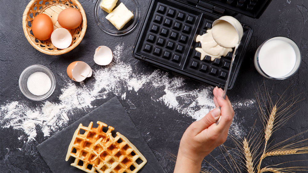 Nalévání těsta na vafle do vaflovače, vedle kterého jsou různé ingredience a již hotové vafle_02