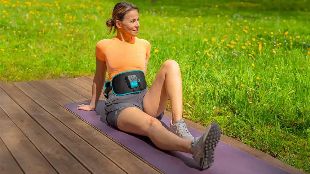 Usmívající se žena sedí na louce a má nasazený elektrostimulátor Beurer Corefit I_03