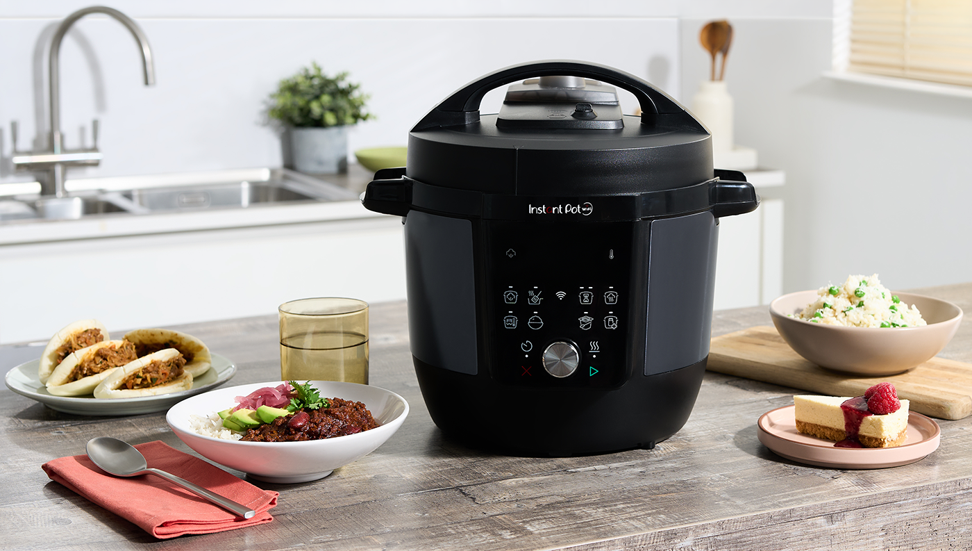 Pohled zepředu na Multifunkční hrnec Instant Pot Plus WiFi 5,7 l stojící na lince s plnými talíři s jídlem