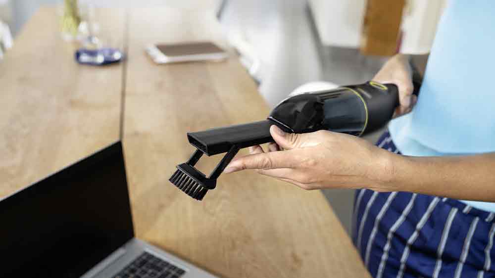 Žena manipulující s ručním vysavačem KÄRCHER CVH Anniversary Edition se štěrbinovým nástavcem s kartáčkem nad stolem s notebookem_03