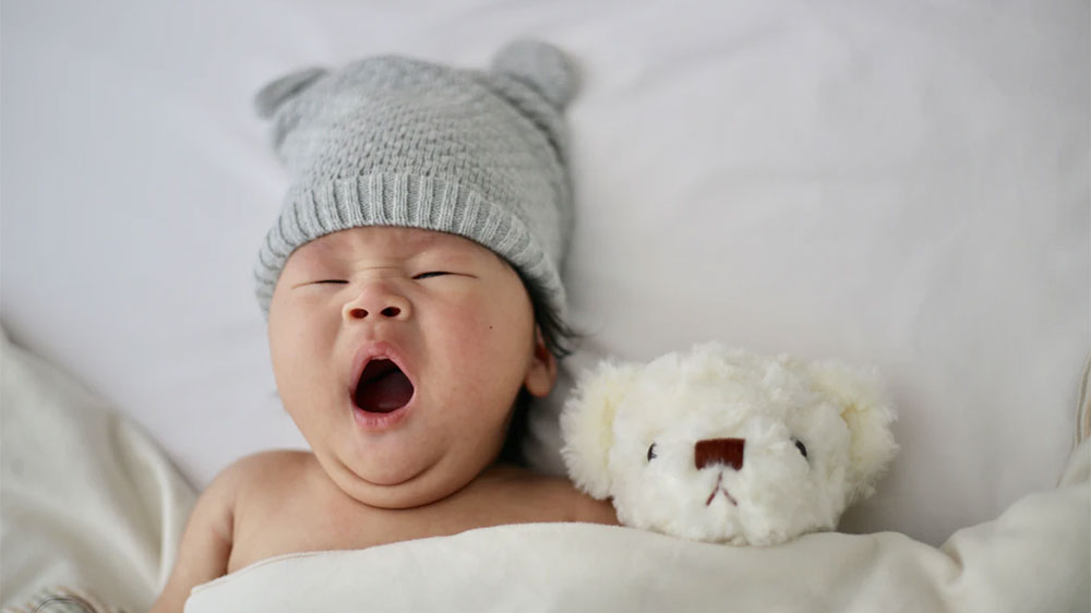 Zívající kojenec v čepičce leží s medvídkem v dětské postýlce