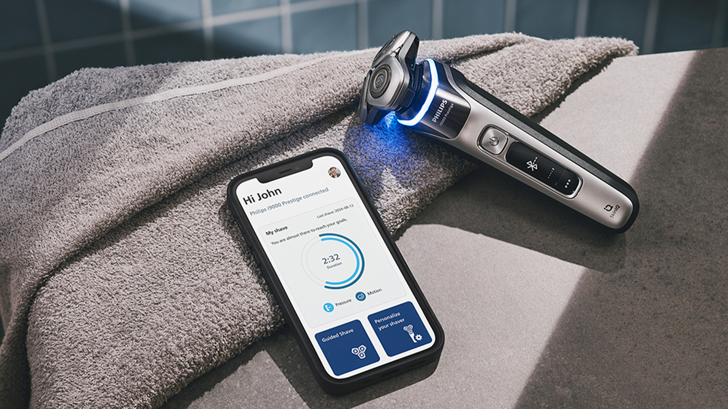 Pohled na Pánský elektrický holicí strojek XP9202/10 Philips Series i9000 Prestige vedle telefonu se zapnutou mobilní aplikací GroomTribe