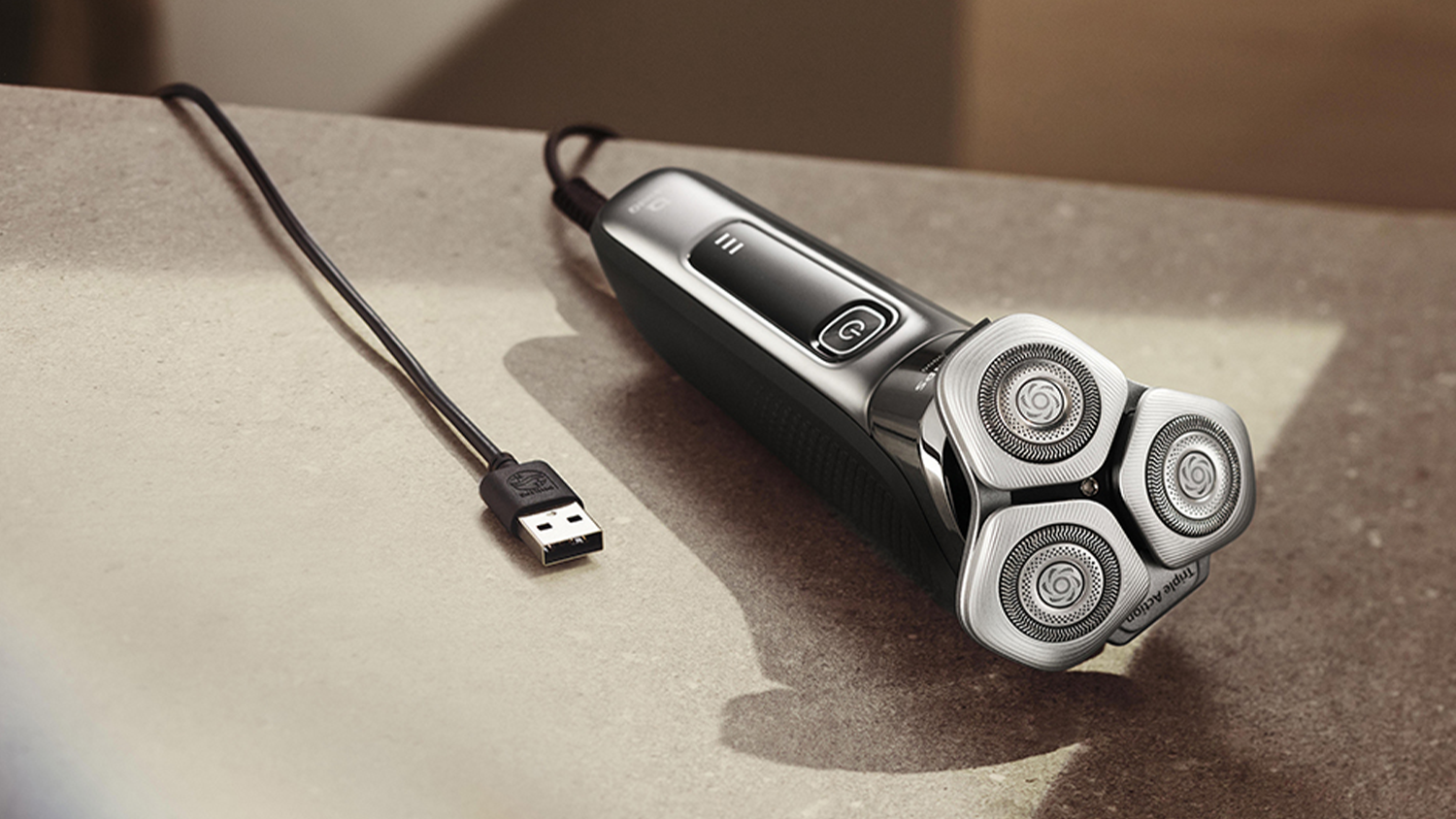 Pánský elektrický holicí strojek Philips Series i9000 ležící na stole s nabíjecím USB kabelem