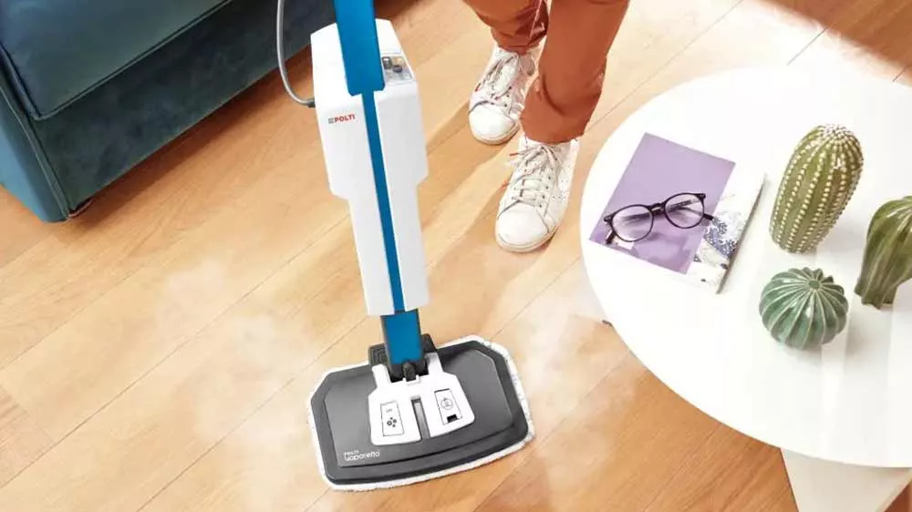 Parní mop Polti Vaporetto SV620_Style stojící na koberci uprostřed obývacího pokoje a dveřmi je vidět prostor kuchyně s mužem u květiny na poličce_02
