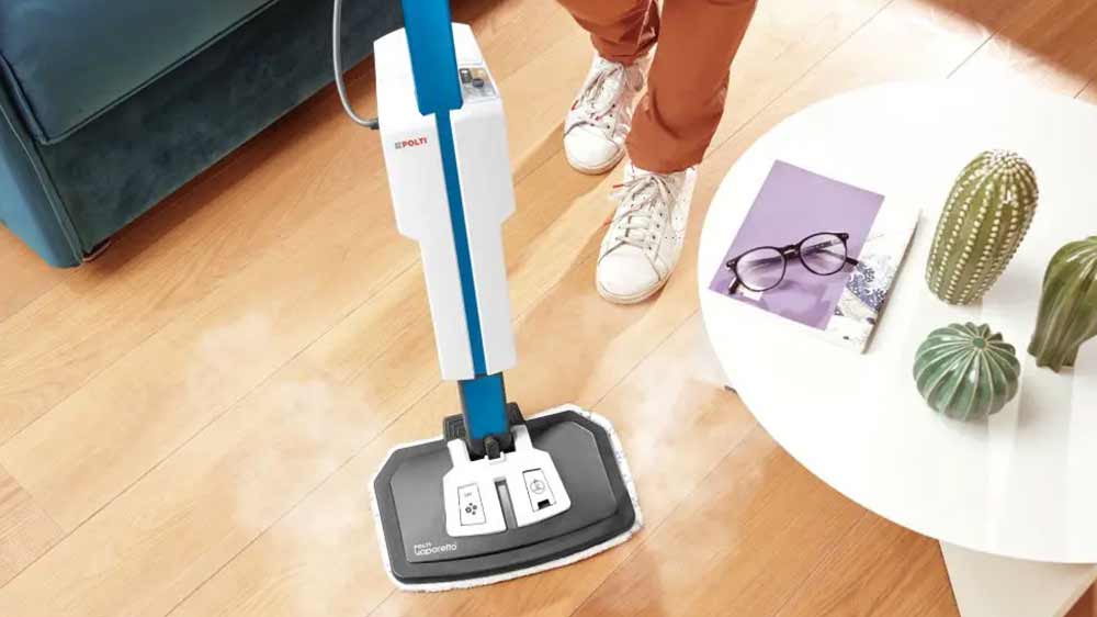Parní mop Polti Vaporetto SV660_Style stojící na koberci uprostřed obývacího pokoje a dveřmi je vidět prostor kuchyně s mužem u květiny na poličce_02