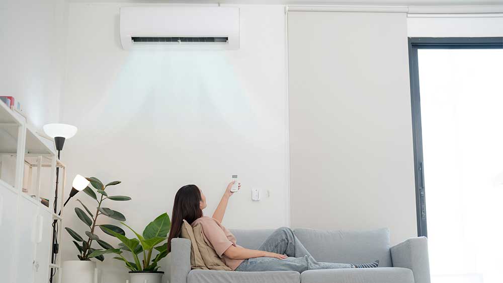 Žena sedící na gauči ovládající nástěnný ventilátor nad sebou dálkovým ovládáním_02