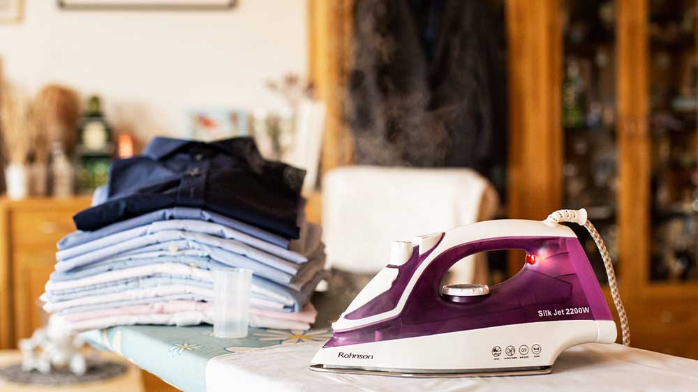 Napařovací žehlička Rohnson R-332 na žehlicím prkně vedle hromádky vyžehleného prádla_02