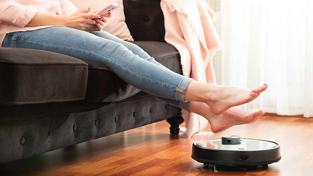 Žena s mobilním telefonem v ruce sedící na gauči se zvednutými nohami, aby uvolnila cestu robotickému vysavači_02