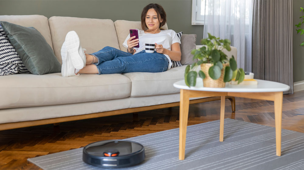 Robotick&yacute; vysavač vys&aacute;vaj&iacute;c&iacute; koberec před pohovkou, na kter&eacute;m lež&iacute; žena s hrnkem n&aacute;poje v ruce a sleduj&iacute;c&iacute; mobiln&iacute; telefon_02