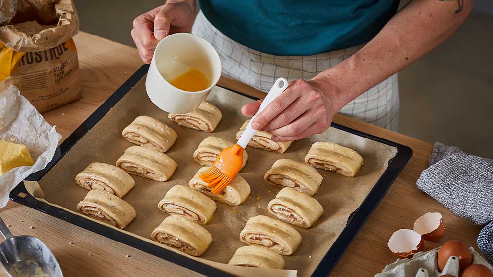 Osoba potírá mašlovačkou nakrájenou roládu na plechu, potírá ji vejcem z hrníčku_02