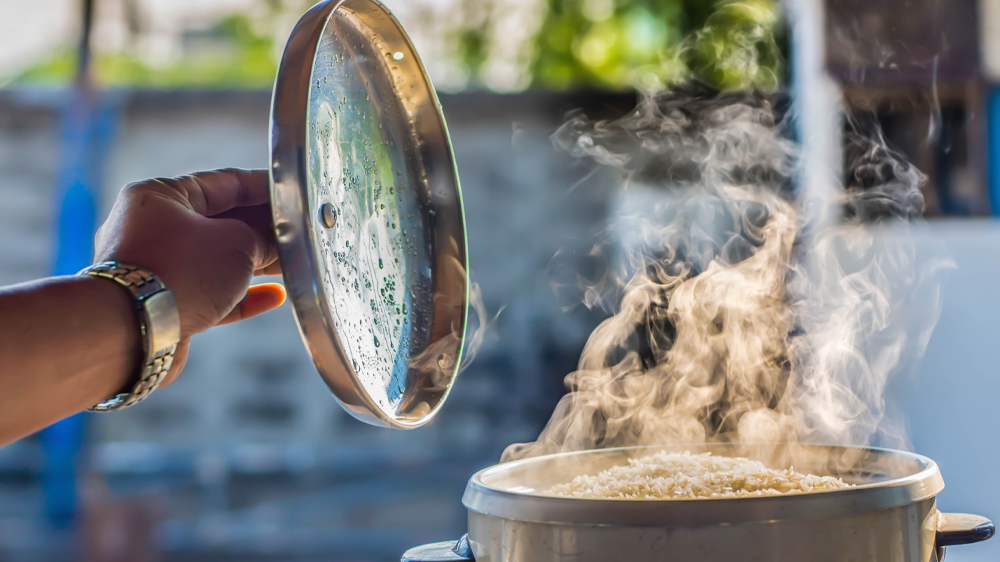Ruka zvedaj&iacute;c&iacute; pokličku z hrnce pln&eacute;ho r&yacute;že, ze kter&eacute;ho unik&aacute; p&aacute;ra_02