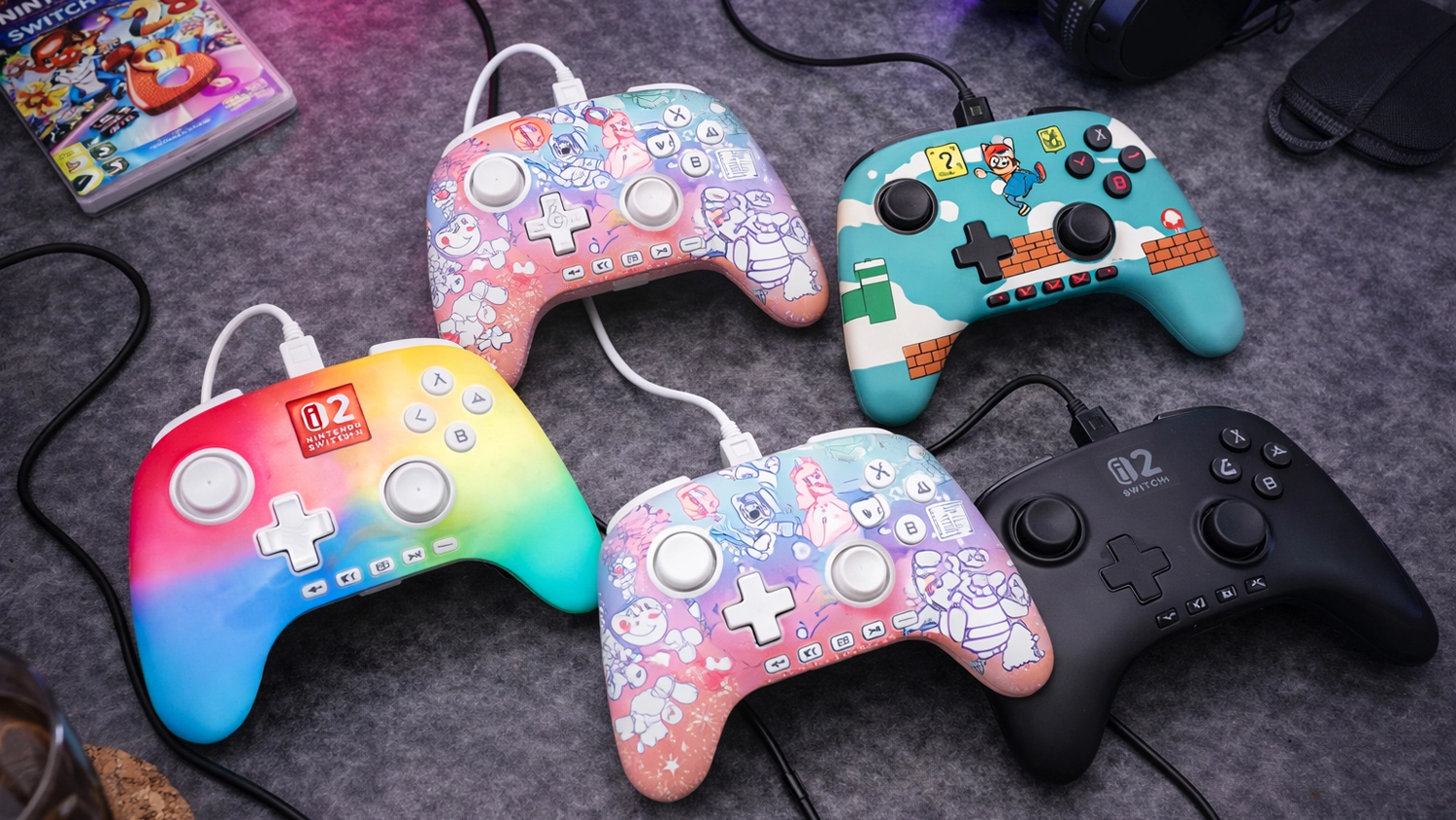 Gamepad PowerA Advantage Wired controller NS2 ve 4 barevných provedeních na herním stole v pozadí s hrou na Nintendo