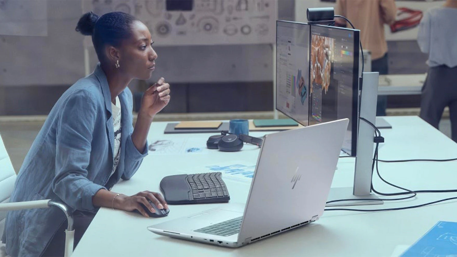 Žena v kancel&aacute;ři u stolu s monitory a HP notebookem