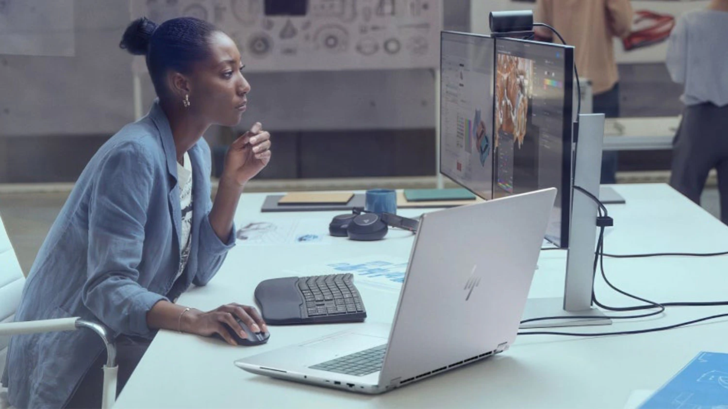 Žena v kancel&aacute;ři u stolu s monitory a HP notebookem